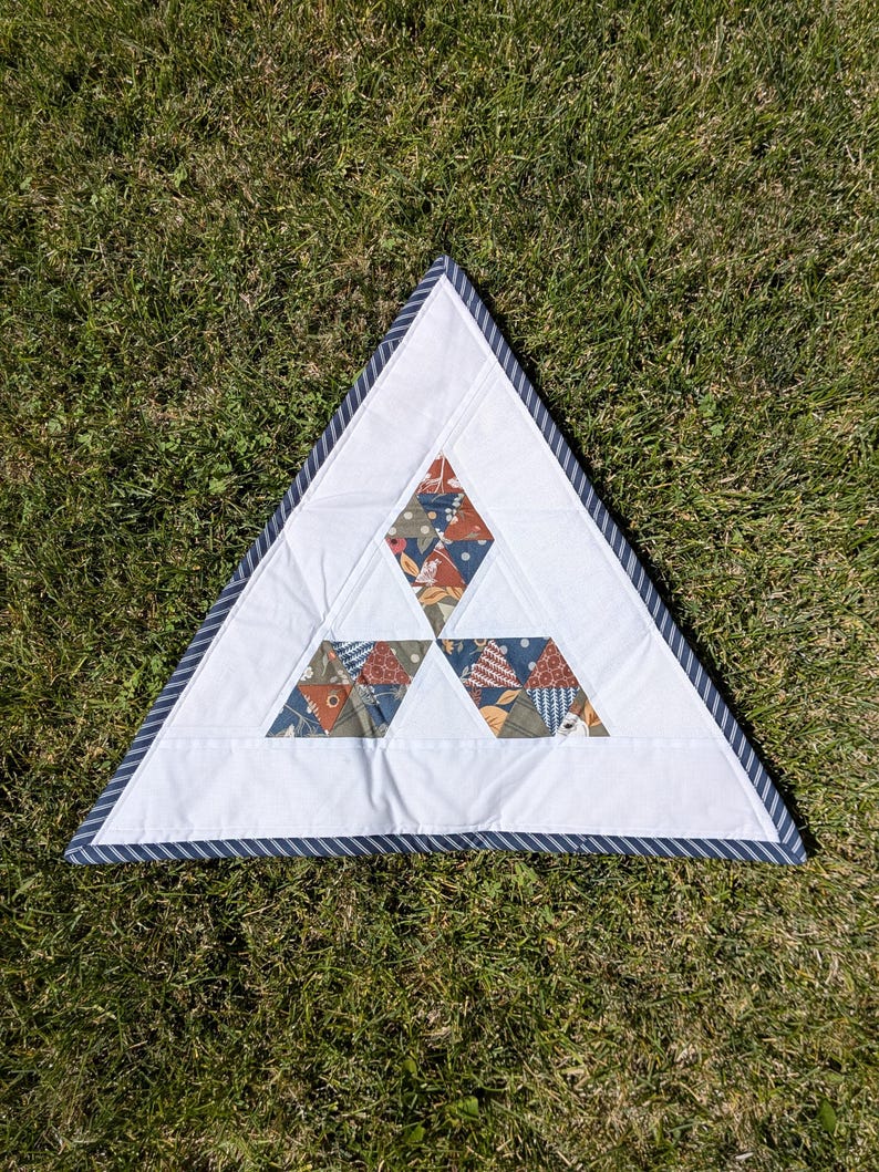 May include: A triangular white quilt with a blue and white striped border. The quilt features three diamond-shaped patches with floral and geometric patterns in shades of orange, blue, and green. The quilt is placed on a bed of green grass.
