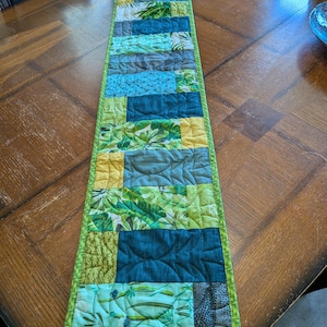 May include: A rectangular table runner with a patchwork design. The runner features various shades of green, blue, and yellow fabrics with leaf and abstract patterns. The edges are trimmed with a solid green border. The runner is approximately 60 inches long.