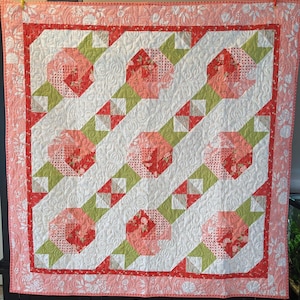 Könnte beinhalten: Eine handgefertigte Steppdecke mit floralem Muster. Die Steppdecke zeigt große, stilisierte Blumen in Rot- und Rosatönen vor einem weißen Hintergrund. Grüne und weiße geometrische Formen und ein rosa Blumenrand vervollständigen das Design.