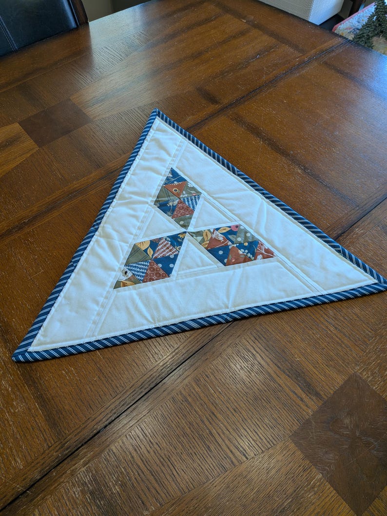 May include: A triangular quilted placemat with a white center and a blue and white striped border. The center features geometric patchwork in shades of blue, green, and red. The placemat is designed for decorative use.