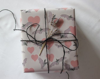May include: A gift wrapped in white paper with a pink and gray bat and heart pattern. The gift is tied with black twine and decorated with dried lavender sprigs.