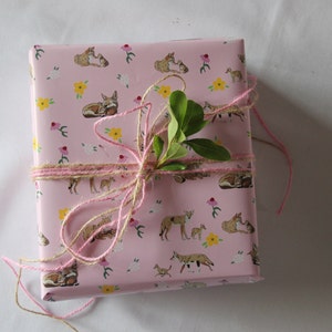 Op de afbeelding: Een roze cadeaudoos ingepakt in papier met een patroon van vossen en bloemen. De doos is vastgebonden met een roze en bruine touw strik en een takje groene bladeren.