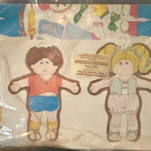 May include: A package of Cabbage Patch Kids paper playmates gift wrap featuring two illustrated doll figures. The left figure wears a red shirt and blue shorts, while the right figure has blonde pigtails and wears a blue vest and pink shorts.