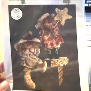 Peut inclure: Un paquet de patrons de poupées avec le texte "PATTERN GARDEN" et "SHAYLA JOY & LADY BUG - 12". L'image présente deux poupées, dont une tenant une baguette en forme d'étoile. Le paquet comprend un transfert de visage. Les poupées sont vêtues de vêtements de style vintage.
