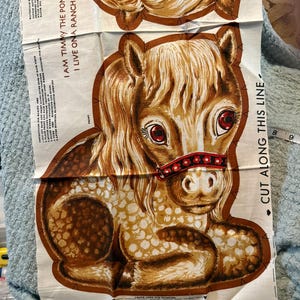 May include: A fabric panel for a stuffed pony toy featuring a brown and white spotted horse with red eyes and a red floral halter. The text reads I AM TIMMY THE PONY I LIVE ONA RANCH and CUT ALONG THIS LINE with sewing instructions printed on the side.