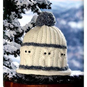 May include: A cream-colored knitted hat with a gray pom-pom on top. The hat features three owl faces with black eyes and a gray band around the bottom and middle. The background shows snow-covered branches and a blurred winter landscape.