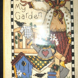 May include: A cross-stitch embroidery featuring a scarecrow wearing a straw hat, a blue birdhouse, a watering can with a heart, and three black crows. The text "I Love My Garden" is stitched in black thread, framed by a black and white checkered border.