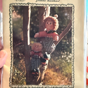 May include: A vintage craft pattern titled "Tomorrow's Treasures" featuring two rag dolls climbing a tree branch. The dolls have curly red hair, striped pants, and are wearing red and green outfits. The pattern is designed by Joanne Burkhart and the name "Clancy" is visible.
