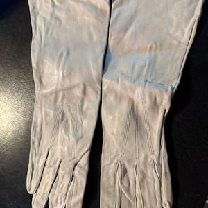 May include: A pair of long, off-white leather gloves featuring three vertical stitched lines on the back of each hand. The gloves extend past the wrist and have a smooth, matte finish with visible natural creasing throughout the material.