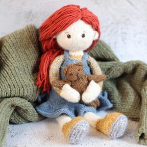 May include: A handmade knitted doll with red yarn hair, wearing a blue dress and holding a small brown teddy bear. The doll has white arms and legs with yellow and grey shoes. A green knitted blanket is in the background.