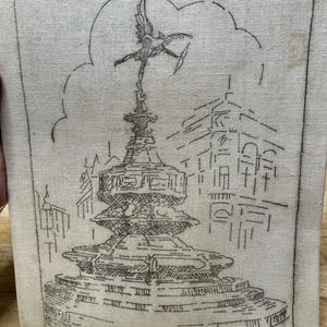 May include: A framed artwork featuring a sketch of the Eros statue in Piccadilly Circus, London. The black line drawing is on a beige fabric background. The artwork includes the text "Eros Statue Piccadilly Circus".