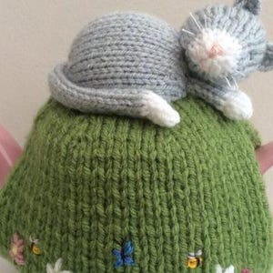 Könnte beinhalten: Eine handgefertigte Teewärmer mit einer gestrickten grünen Hülle, die mit gestickten Blumen und Bienen verziert ist. Eine graue gestrickte Katze liegt obenauf. Die Teekanne hat einen rosa Griff und Ausguss.