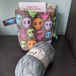 May include: A black drawstring bag with a skull pattern and a green drawstring. The bag is filled with yarn and knitting needles. The bag is sitting on a dark wooden surface. The yarn is a grey and white colour and is labelled "Opal" with a German word for "sock yarn".