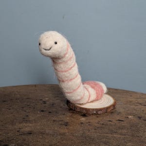 May include: A handcrafted felt worm toy in cream and pink. The worm has a smiling face and is mounted on a small, round wooden base. The body is segmented with pink stripes. A charming, soft toy, perfect for children or as a decorative item.