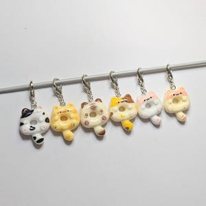 May include: Six cat-shaped charms with a donut design and silver clasps. The charms are in various colours: white, black, yellow, brown, and pink. They are arranged in a row against a white background. Each charm is a unique design.