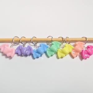May include: A selection of bat-shaped charms in various colours: pink, purple, blue, green, yellow, peach, and magenta. Each charm is attached to a silver keyring and displayed on a wooden dowel against a white backdrop.