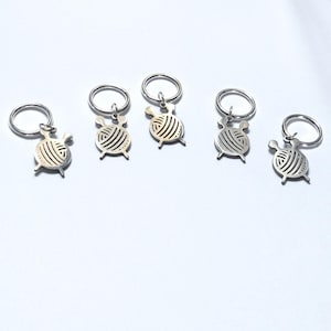 May include: Five silver keyrings, each with a charm shaped like a ball of yarn. The yarn ball design has curved lines to represent the yarn. The keyrings are made of metal and have a circular loop.