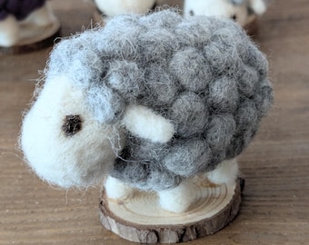 May include: A gray and white felt sheep figurine with a wooden base. The sheep has a fluffy, gray wool body and a white face with black eyes.