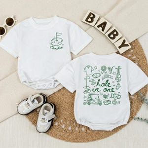May include: White baby bodysuit with a green golf-themed design. One bodysuit has a golf flag and the name "Camden". The other bodysuit has a "hole in one" graphic. Includes white and black baby shoes and sunglasses.