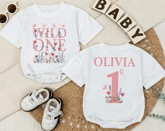May include: Two white baby onesies with floral and butterfly designs. One says "WILD ONE" and the other says "OLIVIA 1". The onesies are next to a pair of white and black baby shoes and wooden blocks that spell "BABY".