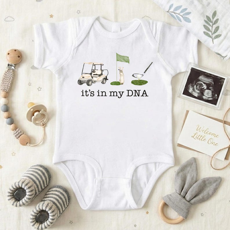 May include: White baby onesie with a golf-themed graphic and the text "it's in my DNA." The design includes a golf cart, flag, and golf club. The image also includes baby accessories like a pacifier, booties, and a teething ring.