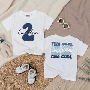 May include: Two white toddler t-shirts. One shirt has the name "Camden" with a navy blue number "2". The other shirt says "TWO COOL" in a repeating pattern of blue and light blue text. Small white sneakers and sunglasses are also in the image.