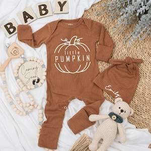 May include: A brown ribbed baby onesie with a white outline of a pumpkin and the text "little PUMPKIN". A brown ribbed hat with the name "Emily" embroidered on it. A crocheted teddy bear with a green ribbon around its neck.