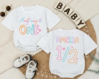 May include: Two white baby onesies with colorful text. One says "Half way to ONE" and the other says "AMELIA 1/2". The image also includes a pair of white baby shoes and wooden blocks spelling "BABY".