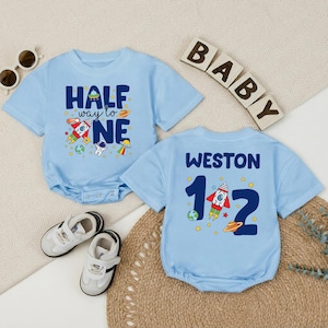 May include: Two light blue baby onesies with space-themed graphics. One says "HALF way to ONE" with rockets and planets. The other says "WESTON 1 1/2" with a rocket and planets. Includes baby shoes and sunglasses.