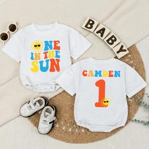Könnte beinhalten: Zwei weiße Baby-Bodys mit farbenfrohem Text und Grafiken. Einer sagt "ONE IN THE SUN" mit einer Sonnengrafik. Der andere sagt "CAMDEN 1" mit einer Sonnengrafik. Außerdem ein Paar weiße Babyschuhe und Sonnenbrillen.