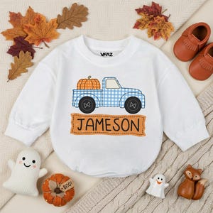 May include: A white long-sleeve sweatshirt featuring a graphic of a blue and white checkered pickup truck carrying an orange pumpkin. Below the truck is an orange banner with the name JAMESON printed in black. The brand VFAZ is on the collar.