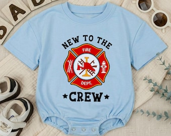May include: A pale blue baby bodysuit featuring a firefighter emblem. The emblem is red, yellow, and white, with the text "NEW TO THE FIRE DEPT. CREW". The bodysuit has short sleeves and popper fastenings.