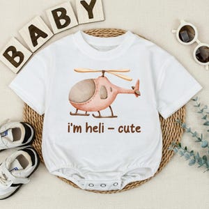 May include: White baby onesie with a watercolor illustration of a pink helicopter. The text "i'm heli - cute" is printed in brown below the helicopter. The onesie has short sleeves and a snap closure.