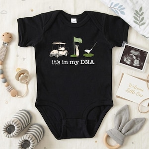 May include: Black baby onesie with a golf-themed graphic. The design includes a golf cart, flag, golf club, and the text "it's in my DNA." The onesie is surrounded by baby accessories, including a pacifier, rattle, and ultrasound photo.