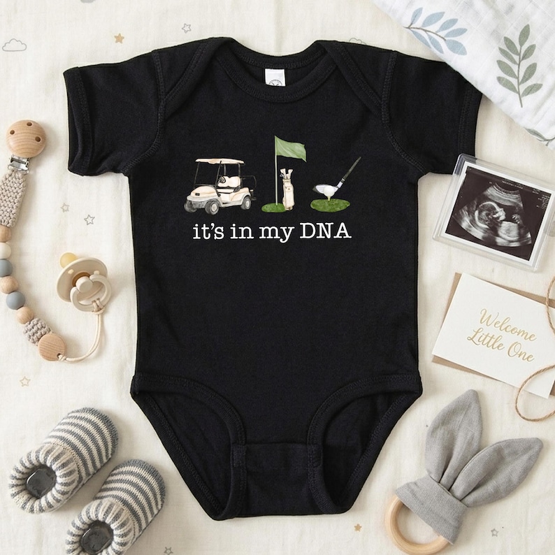 May include: Black baby onesie with a golf-themed graphic. The design includes a golf cart, flag, golf club, and the text "it's in my DNA." The onesie is surrounded by baby accessories, including a pacifier, rattle, and ultrasound photo.