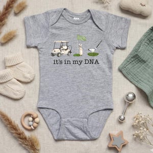 May include: A gray baby onesie with a golf-themed graphic. The design includes a golf cart, a flag, a golf club, and the text "it's in my DNA". The onesie is surrounded by baby accessories, including socks, a rattle, and a wooden toy.