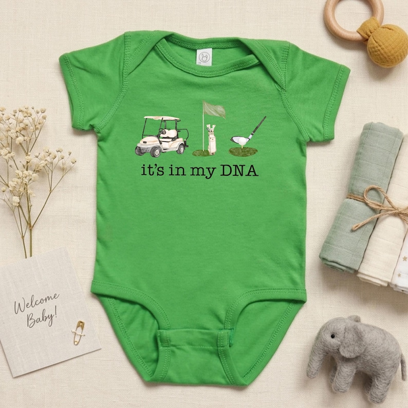 May include: Green baby onesie with a golf-themed graphic. The design includes a golf cart, a flag, a golf bag, and a golf club hitting a ball. The text "it's in my DNA" is printed below the graphic.