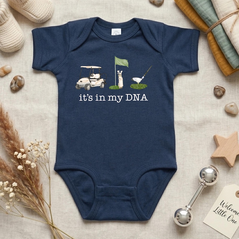 May include: Navy blue baby onesie with a golf-themed graphic. The design includes a golf cart, a flag, a golfer, and a golf ball. The text "it's in my DNA" is printed below the illustrations. A perfect gift for a baby.