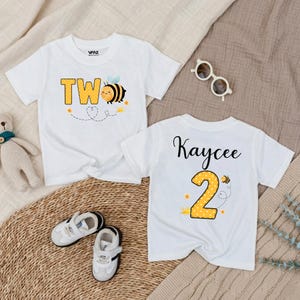 May include: Two white toddler t-shirts with bee-themed designs. One shirt features the word "TWO" in yellow block letters with a bee graphic. The other shirt has the name "Kaycee" and the number "2" with a bee graphic. Small white sneakers are next to the shirts.