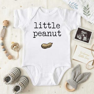 May include: White baby onesie with the text "little peanut" and a peanut graphic. The image includes baby accessories like a pacifier, booties, and a bunny ear teether. A sonogram and a card that says "Welcome Little One" are also included.