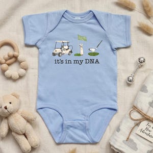 May include: Light blue baby onesie with a golf-themed graphic. The design includes a golf cart, a flag, a golf club, and the text "it's in my DNA". The onesie is surrounded by baby accessories.