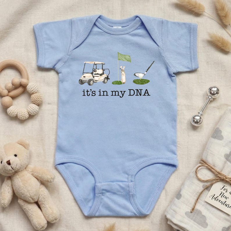 May include: Light blue baby onesie with a golf-themed graphic. The design includes a golf cart, a flag, a golf club, and the text "it's in my DNA". The onesie is surrounded by baby accessories.