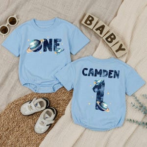 May include: Two light blue short-sleeved rompers featuring space-themed graphics. One romper displays the word ONE with a planet and rocket. The other displays the name CAMDEN, the number 1, a rocket, and a planet. Both have snap closures.