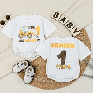 May include: Two white baby onesies with a construction theme. One says "I'M 1 AND DIGGING IT" with a yellow excavator. The other says "CAMDEN 1" with a brown number one and a yellow excavator. Includes baby shoes and sunglasses.