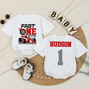 May include: Two white short-sleeved baby bodysuits. One features a red race car graphic with the text FAST ONE and checkered flags. The other features the name HUDSON in red letters above a large number 1 with a black and white checkered pattern.