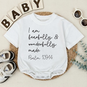 May include: A white short-sleeved baby bodysuit featuring black text that reads I am fearfully & wonderfully made Psalm 139:14. The garment has a round neckline and a snap-button closure at the bottom, presented with white sneakers and sunglasses.