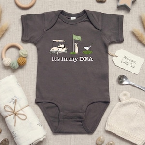 May include: Gray baby onesie with a golf-themed graphic. The design includes a golf cart, flag, and golfer, with the text "it's in my DNA." A tag reads "Welcome, Little One."