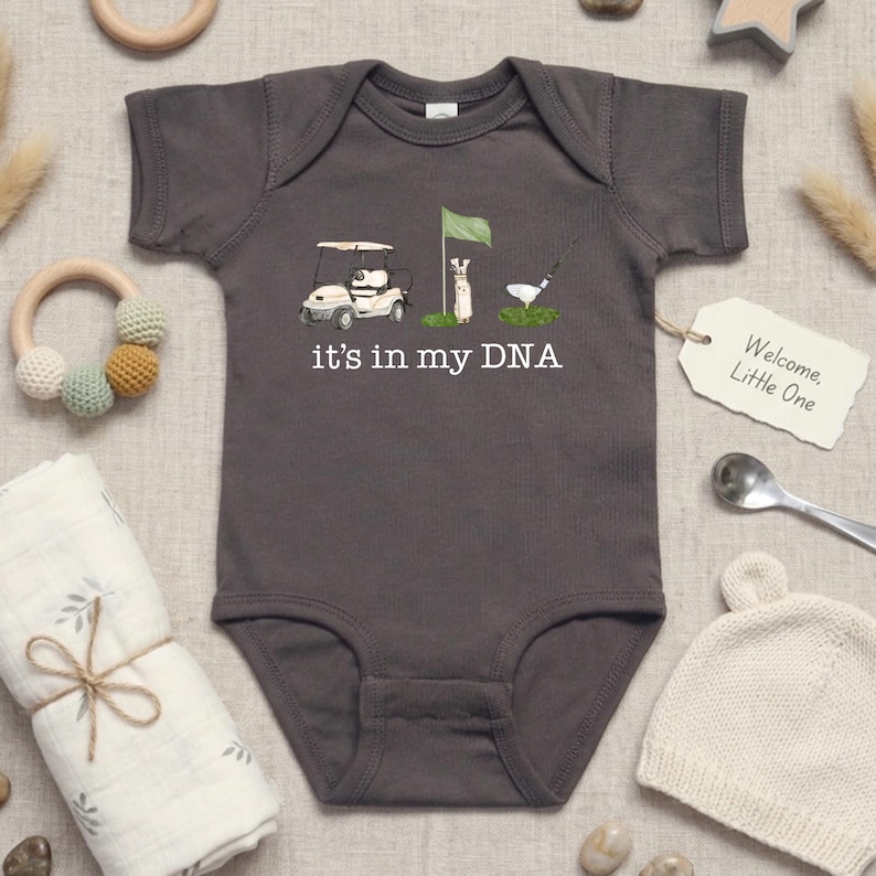May include: Gray baby onesie with a golf-themed graphic. The design includes a golf cart, flag, and golfer, with the text "it's in my DNA." A tag reads "Welcome, Little One."