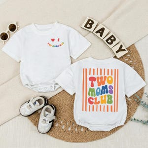 May include: Two white short-sleeved baby bodysuits. One features a small chest graphic with a heart, lightning bolt, and the text "Two Moms Club." The other displays a large back graphic with orange vertical stripes and the text "Two Moms Club" in colorful, wavy, retro-style lettering.