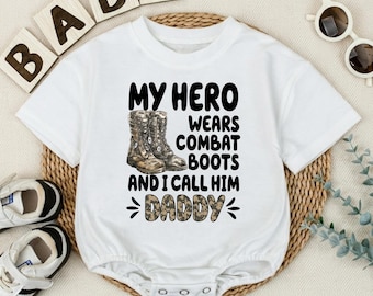 May include: A white short-sleeved baby bodysuit featuring a graphic print of two brown combat boots. The text reads: MY HERO WEARS COMBAT BOOTS AND I CALL HIM DADDY, with the word DADDY displayed in a camouflage pattern and black lettering.